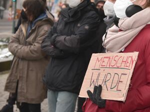 Draußen. Winter. Menschen mit FFP2 Masken in warmer Kleidung. Ein Mensch mit roter Jacke, schwarzen Handschuhen, und hellem Schal hält ein Pappschild vor die Brust. TextMENSCHENWÜRDE