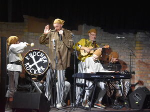 Schauspieler Jung und alt, an Trommel, Keybord und Mikrofon. Auf der Trommel sind gekreuzte Knochen abgebildet und KNOCHEN-ROCK steht drauf. Die Bandmitglieder tragen schwarz-weiß-gestreifte Hosen, langärmlige Hemden und braune Kopftücher