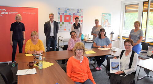 Menschen in einem Raum. Fünf Personen stehen und fünf sitzen im Raum verteilt. Alle sehen in die Kamera.