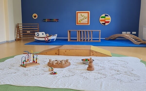 Im Vordergrund: Decke mit Spielzeug für Babys. Im Hintergrund Kletterelemente und eine blaue Wand mit Bild.