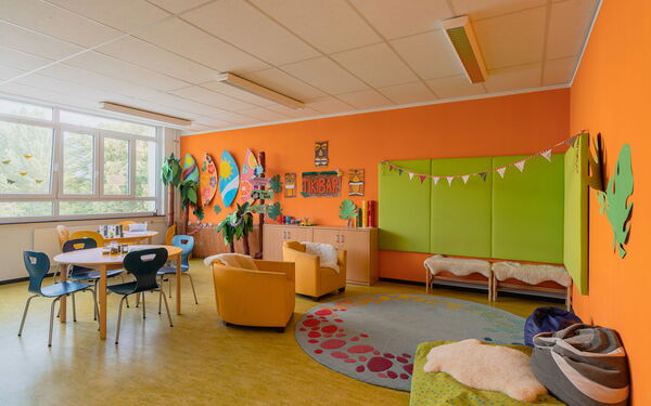 Raum mit orangene Wänden, lichtspendenen Fenster, im Raum ein großer runder Teppisch, zwei gelbe Sessel, zwei Tische mit je 4 Stühlen vor den Fenstern. Gebastelte Wanddekoration.
