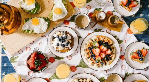 Frühstückstisch von oben mit Tellern voller Waffeln, Beeren und Gebäck, dazu Tassen und Gläser mit Getränken.
