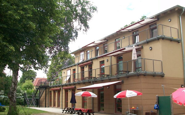 Dreistöckiges Kitagebäude. Im ersten und zweiten Stock Terrassenähnliche Außenbereiche. Vor dem Gebäude stehen Tische, Bänke und 2 Sonnenschirme.