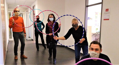 Vier Personen in einem Flur. Alle tragen Stoffmasken. Alle halten bunte Hula Hoop Reifen hoch.