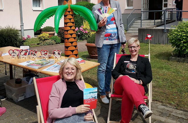 Ein Haus, im Vordergrund zwei Frauen sitzen in roten Sonnenstühlen unter einer aufblasbaren Gummipalme. Eine Frau steht hinter ihnen mit einem Fächer in der Hand.