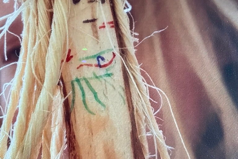 Basteln mit Naturmaterialien. Länglicher Ast mit abgeschabter Rinde, darauf eine farbige abstrakte Zeichnung grün, blau und rot. Der Ast ist mit mehreren losen, beigefarbenen Fäden umwickelt.
