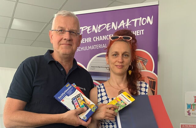 Mann und Frau stehen nebeneinander im Büro. Beide halten Schulmaterial in den Händen, Hefter, Stifte, Tuschkasten. Mann hat graue kurze Haare, Brille und ein dunkelblaues Poloshirt. Frau hat lange rote Haare, Brille auf den Kopf geschoben in trägt Kleid.