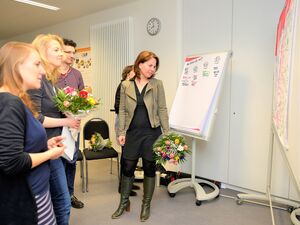 Frau mit Blumenstrauß steht vor einem Flipchart. Eine weitere Frau hat einen Bumenstrauß in der Hand. Die 4 Personen schauen interessier auf eine Tafel mit Notizen.
