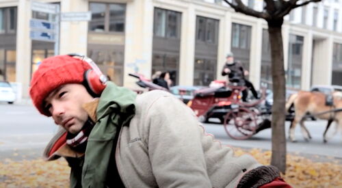 Draußen in einer Stadt. Ein Mann mit roter Mütze und roten Kopfhörern, graue Jacke. Im Hintergrund eine Pferdekutsche.