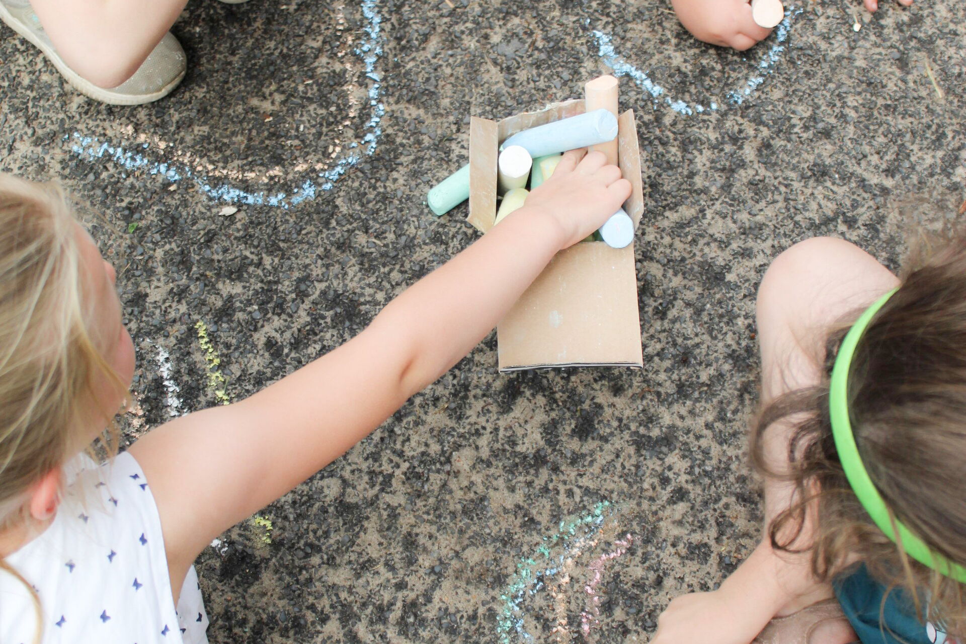 4 kleine Kinder sitzen / hocken am Boden auf Stein. In der Mitte ein kleiner Karton mit Kreide. Sie malen mit Kreide.