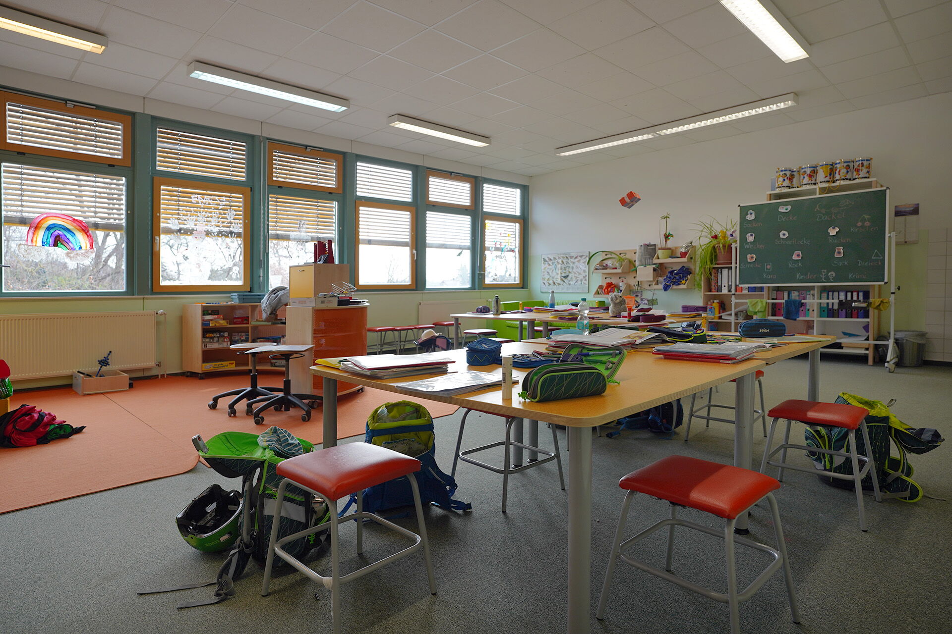 Lernzimmers. großer Tisch in der Mitte des Raumes, darauf Federtaschen, Blöcke und Hefter. Um den Tisch stehen Hocker und Schulranzen.Vorne eine grüne Schultafel.