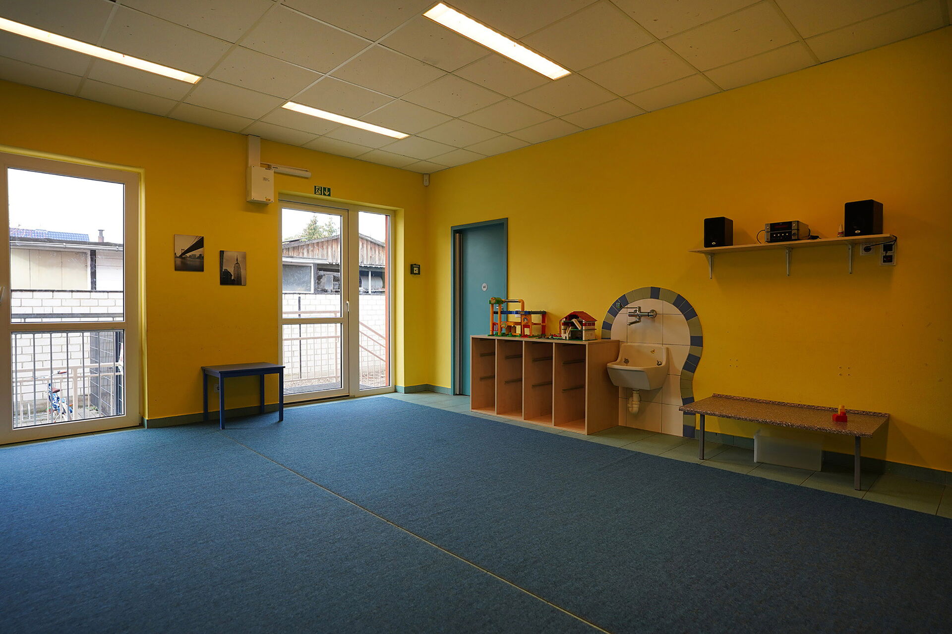 Großer Raum, gelbe Wände, Teppich blau. An der Wand ein Regalbrett wo eine Musikanlage mit zwei Boxen steht.