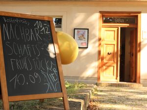 Draußen vor einer geöffneten Tür steht ein Schild mit einem gelben Luftballon. Über der Tür steht die Hausnummer 98a. Auf dem Schild steht: kostenfreies Nachbarschaftsfrühstück Hier. 10.09. von 10 bis 12 Uhr.