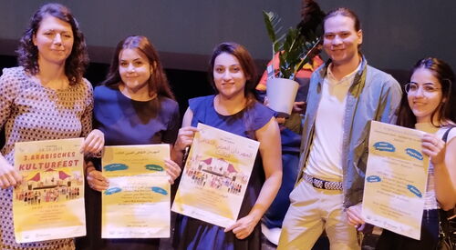 Vier Frauen halten Plakate in die Kamera und lächeln. Von rechts die zweite Person ist ein Mann, der eine Grünpflanze hält und lächelt.