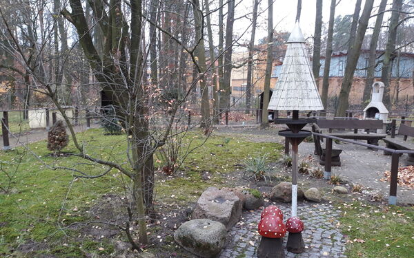 Kleine Parkanlage. Kahle Bäume und Sträucher, im Vordergrund 3 Deko Fliegenpilze, rechts 3 Bänke.