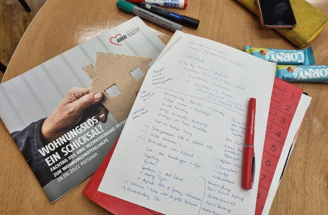Tischplatte darauf liegen Stifte, ein AWO Flyer: Wohnungslos - ein Schicksal. Corny Riegel, Geldbörse und darauf ein Smartphon. DIN A4 Blatt mit handschriftlichen Notizen.
