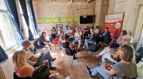 Großer Raum. Ca. 20 Menschen sitzen in einem Raum in mehreren Kreisen.