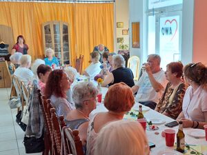 Lange Tafel in einem großen Raum. 16 ältere Menschen sitzen an der langen Tafel und essen und trinken.