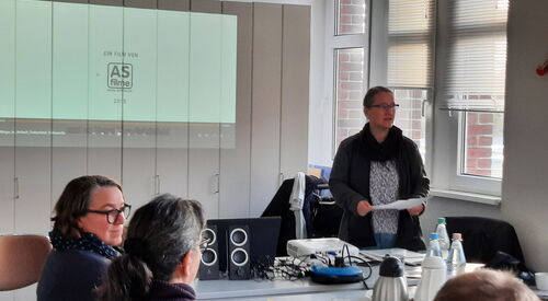 Konferenzraum. Zwei Frauen sitzen am Tisch. Eine Frau steht vorne mit Notizen in den Händen und spricht. Auf dem Tisch sind Getränke und Geschirr.