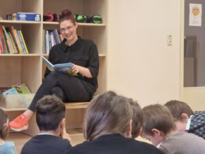 Eine Frau sitzt mit einem Buch in der Hand in einem Raum mit Regalen. Sie lächelt die Kinder, die vor ihr auf dem Boden sitzen an.