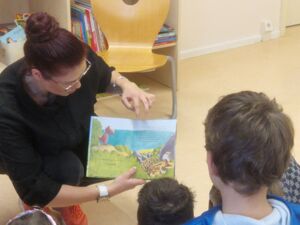 Frau hat sich hingehockt und hält ein Buch aufgeklappt und es den Kindern zu zeigen. Die Frau hat lange Haare zum Dutt gedreht, Brille und schwarze Kleidung.