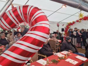 Im inneren des großen Festzeltes. Tische Stühle, viele Menschen. Der Chor steht vorn und singt. Im Vordergrund des Bildes ist ein Luftbalon in Form ein rot-weiß gestreiften Zuckerstange.