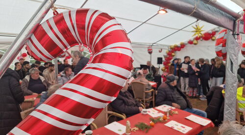 Im inneren des großen Festzeltes. Tische Stühle, viele Menschen. Der Chor steht vorn und singt. Im Vordergrund des Bildes ist ein Luftbalon in Form ein rot-weiß gestreiften Zuckerstange.
