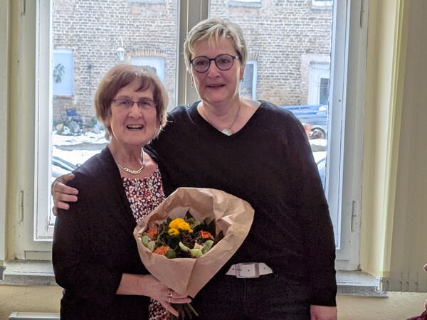 Zwei lächelnde Frauen in schwarzer Kleidung. Die Frau links hält einen Blumenstraß in der Hand.