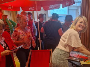 Menschen in einem Raum. Ein roter AWO Sonnenschirm, eine aufblasbare Palme und ein Liegestuhl. Eine Frau mit Brille steht an einem Tisch mit Werbe-Material der AWO und lächelt in die Kamera.