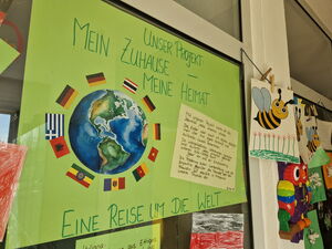 Großes grünes Plakat an einer Glasscheibe. Text: Unser Projekt: Mein Zuhause - Meine Heimat    Bild einer Erde/Welt mit Landesflaggen drum rum.