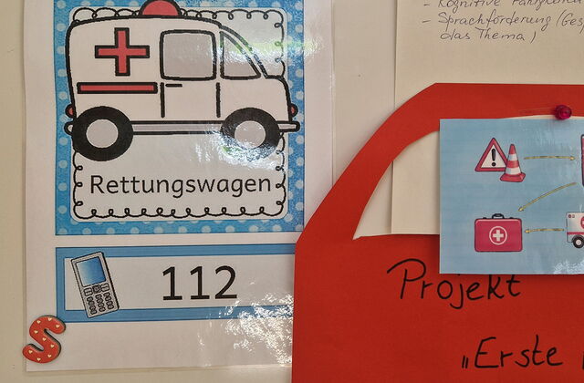 Ein Anschauungsbild von einem Rettungswagen. Darunter ein Telefon und ganz groß die 112. Informationen sind an eine Wand geklebt.