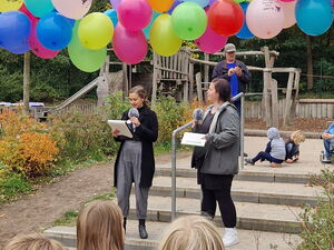 Zwei Frauen stehen auf einer Treppe und halten eine Rede. Über ihnen sind viele farbige Luftballons.