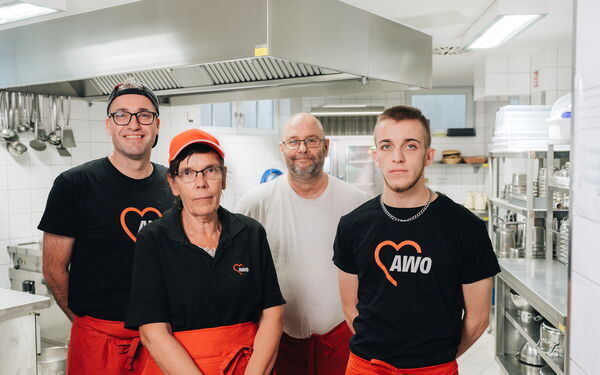 Saubere Küche. Gruppenbild. 4 Personen. 3 Männer und eine Frau. Sie tragen Dienstkleidung und rote Kochschürzen.