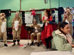 Vier Menschen im Rentierkostüm, eine sitzt und spielt Gitarre dazwischen eine Frau im roten Kleid mit Feenflügeln. Raum ist weihnachtlich dekoriert.