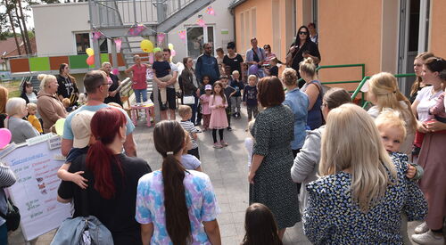 Viele Menschen / Kinder stehen neben einem Gebäude. Sie stehen in einem großen Kreis. Mütter mit kleinen Kindern auf dem Arm. Eine Treppe ist mit Luftballons und Wimpel geschmückt.