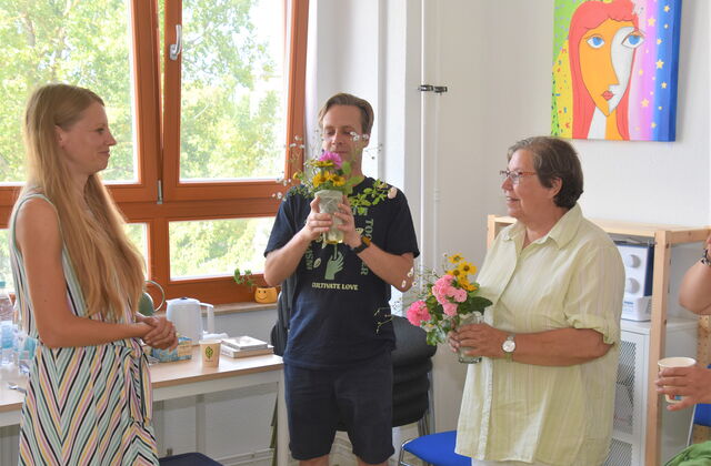 Beide Künstler stehen nebenienander und halten kleine Blumensträuße im Wasserglas in der Hand. Jüngerer Mann in blauem T-Shirt und kurze Hose. Ältere Frau, kurze graue Haare und Brille mit heller Bluse. Freu mit langen blonden Haaren steht ihnen gegenüber