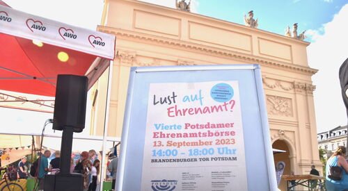 Draußen, schönes, sonniges Wetter. Das Brandenburger Tor in Potsdam im Hintergrund. Ein Aufsteller verkündet die vierte Potsdamer Ehrenamtsbörse am 13.September 2023 14 bis 18 Uhr.