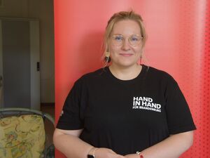 Portrait. Junge Frau mit blonden, langen Haaren, großer runden Brille und Ohrringen. Sie trägt ein schwarzes T-Shirt auf dem in weißer Schrift: "Hand in Hand für Brandenburg" steht.