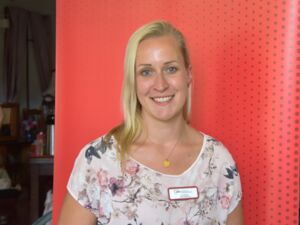 Portrait. Junge Frau mit langen glatten blonden Haaren, die ihr rechts über die Schulter fallen, schmales freundliches Gesicht, strahlendes Lächeln. Sie trägt ein helles T-Shirt mit Pflanzen-Musterung und eine Kette.