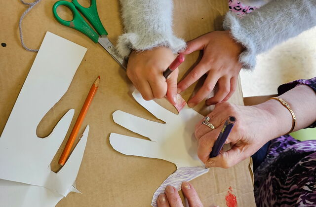 Ein Kind und ein älterer Mensch malen gemeinsam eine Hand aus. Nur die Hände sind zu sehen.
