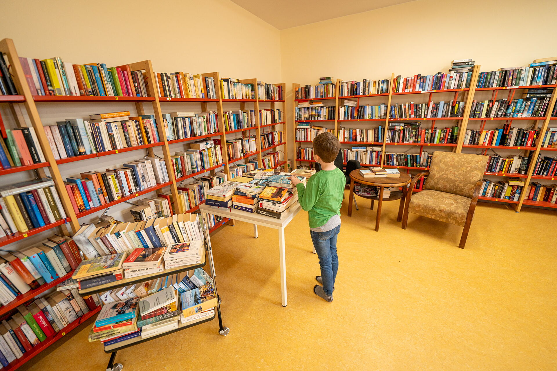 Kind steht an einem Tisch und schaut sich ein Buch an. Bibliothek, Regale mit vielen Büchern stehen an den Wänden.