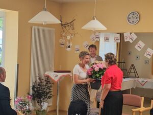 In einem großen Raum mit großen Fenster. Eine Frau überrecht einen Topf mit Blumen an eine Frau im roten AWO Poloshirt und schwarzem Rock. Anwesende sitzen im Raum. Ein Durchgang ist mmit AWO Wimpel geschmückt.