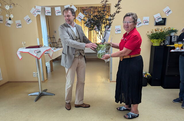 Großer Raum. Ein Stehtisch. Ein Mann übergibt eine Topfpflanze an eine Frau. Der Mann ist groß, krauses graue Haar, graues Jackett, weißes Hemd, helle Hose, braune Schuhe, Frau mit Brille, rotem AWO Poloshirt und schwarzen Rock, in Sandalen.