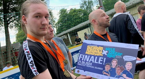 Junger Mann hält den ersten Preis in den Händen, er ist blond und hat eine Medaille an einem orangen Band um den Hals hängen. Preis: große Karte Text: "Straßenfußball. Finale 21.09.2024 Finalfest Tropical Island". Zwei weitere Spieler mit Medaille im Bild
