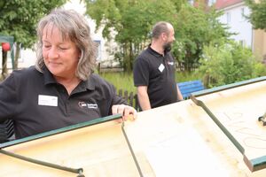 Eine Frau die einen Tisch aufbaut. Ein Mann steht in Hintergrund. Beide tragen schwarze Poloshirts mit AWO Logo Aufdruck.