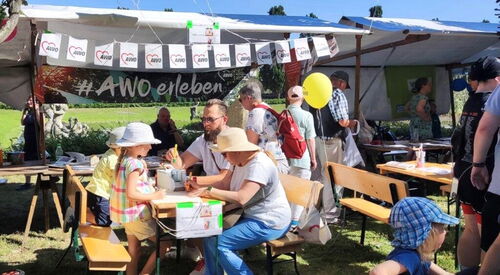 Vor Marktständen sind Tische und Bänke aufgestellt, an denen 2 Kinder und 2 Erwachsene sitzen und die Kinder malen Bilder. Der Stand ist mit Luftballons und Wimpeln geschmückt. Die Sonne scheint.