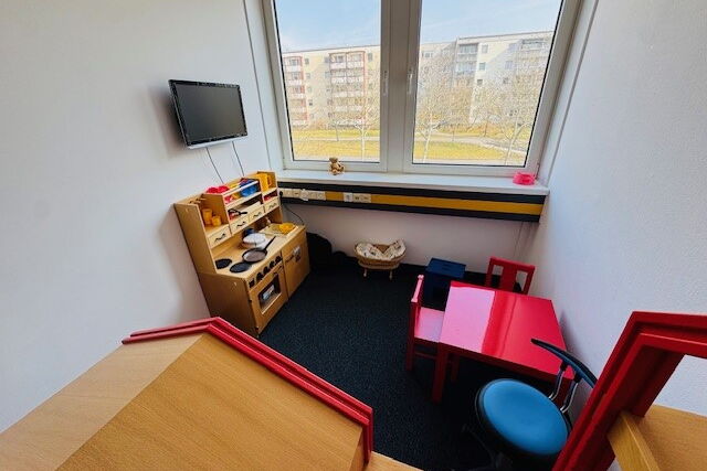 Ein Raum mit ausgestatteter Spielküche. Gegenüber steht ein roter Kindertisch mit zwei Stühlen, unter dem Fenster ein Puppenbett. An der Wand hängt ein Monitor.