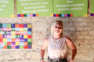 Portrait. Junge Frau. Blonde glatte schulterlange Haare, helles Achselshirt. Sie lächelt freundlich in die Kamera. Sie steht vor einer Steinwand. An der Wand sind Schilder mit Forderungen.