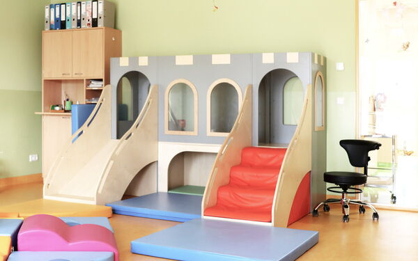 Spielzimmer, Spiellandschaft. Mit roter Treppe auf der rechten Seite und Rutsche auf der linken Seite, sieht aus wie eine Burg. Daneben in der rechen Ecke steht ein hohes Regal mit Schranktüren.