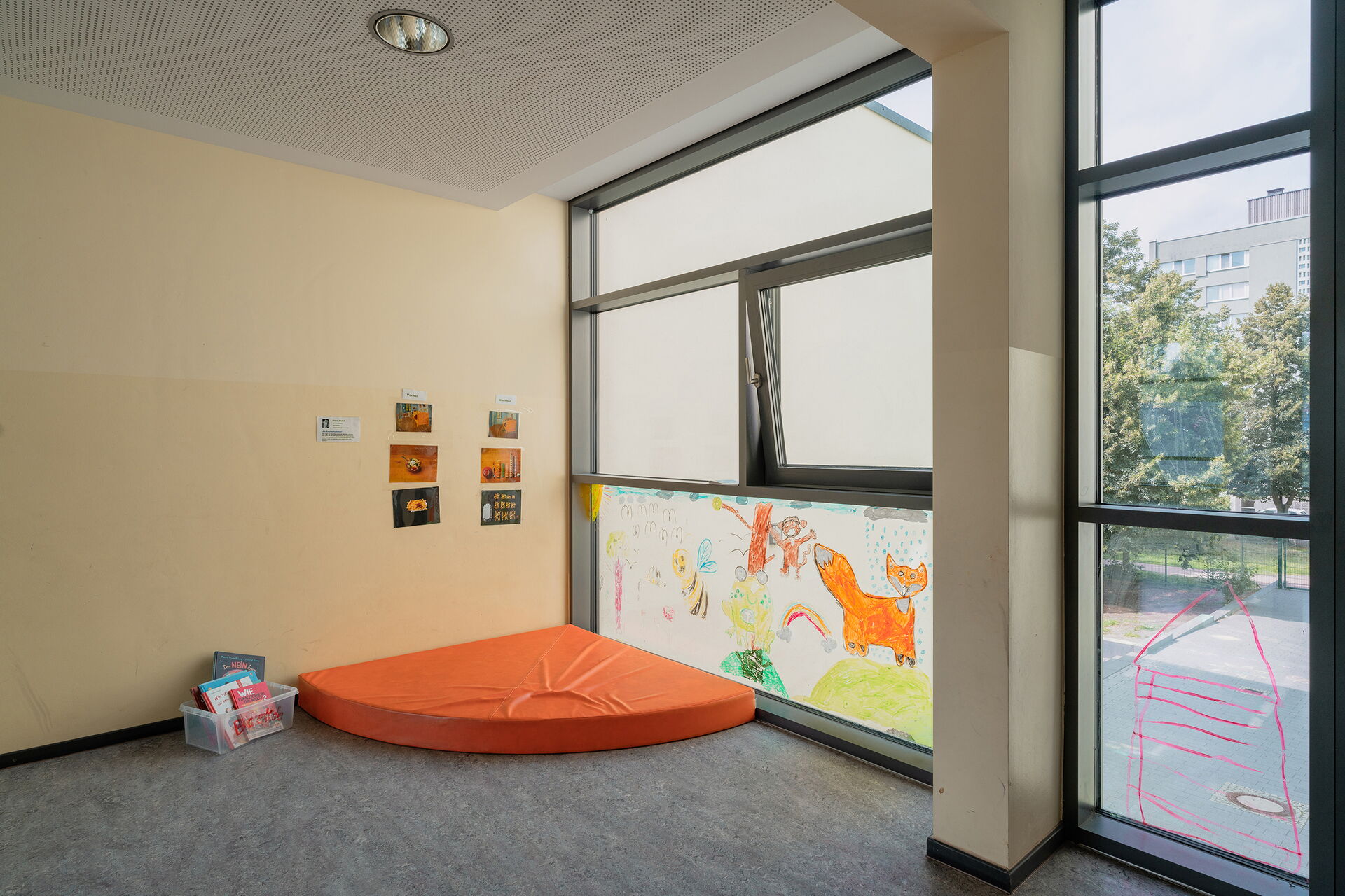 Ecke eines Raums. Eine orangene Matratze liegt in der Ecke, das untere Fenster der Ecke ist bamalt mit Fuchs, Biene, Regenwolke und Sonne.